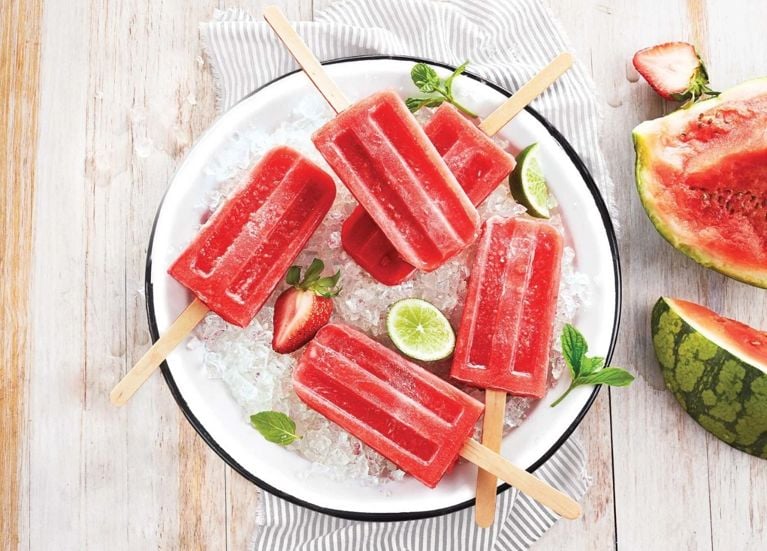 Strawberry-Watermelon Limoncello Ice Pops on a white plate filled with crushed ice next to slices of lime and fresh strawberries and fresh mint on a striped napkin on a wood table next to a cut watermelon and more strawberries