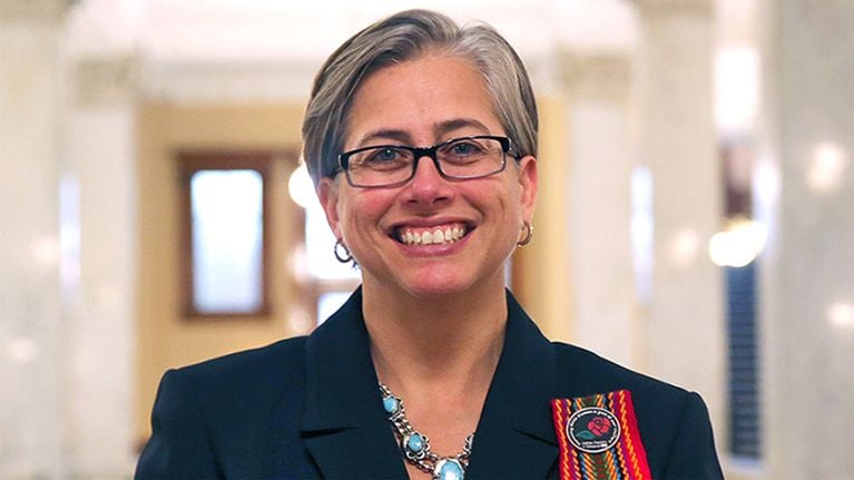 A person with short, grey hair wearing a navy blazer and black-rimmed glasses
