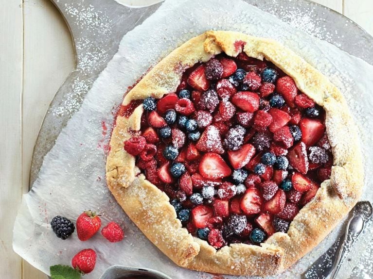 A very berry galette topped with blueberries, raspberries, strawberries and blackberries on a pizza stone next to a small measuring cup full of berries and leaves sprinkled with sugar for an easy, gorgeous, delicious berry galette recipe