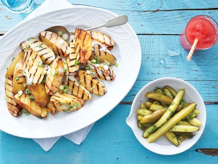 A white platter of warm grilled potato salad topped with green onions next to a plate of dill pickles and a glass of pink watermelon lemonade