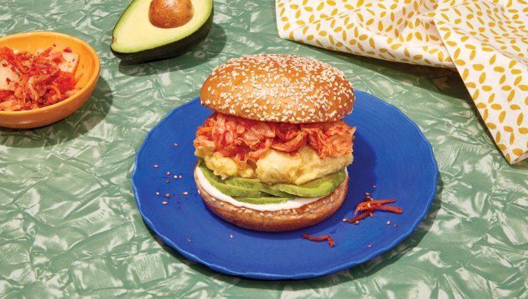 A table with a variety of items: a bowl of kimchi, half an avocado, a printed napkin, and a kimchi, egg and avocado sandwich on a blue plate.