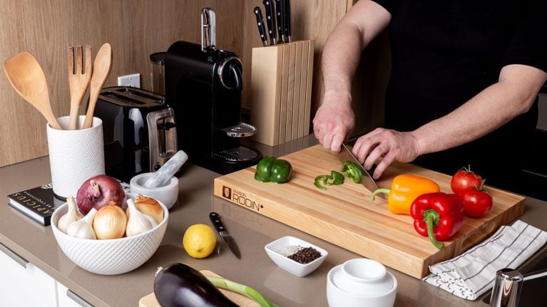 Hands cutting peppers over an edge-grain wooden cutting board surrounded by a counter full of vegetables and kitchen utensils for a post on wood cutting boards.