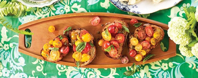 A wood cutting board with four slices of barbecue bruschetta, made with grilled crusty Italian bread and topped with cherry tomatoes and an olive oil and caper vinaigrette as well as fresh basil for a post on an easy grilled bruschetta recipe