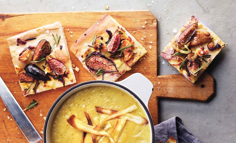Three squares of Fig, onion and roasted garlic focaccia on a cutting board next to a bowl of soup; the focaccia is also topped with goat cheese and rosemary for a post on a Fig, onion and roasted garlic focaccia recipe