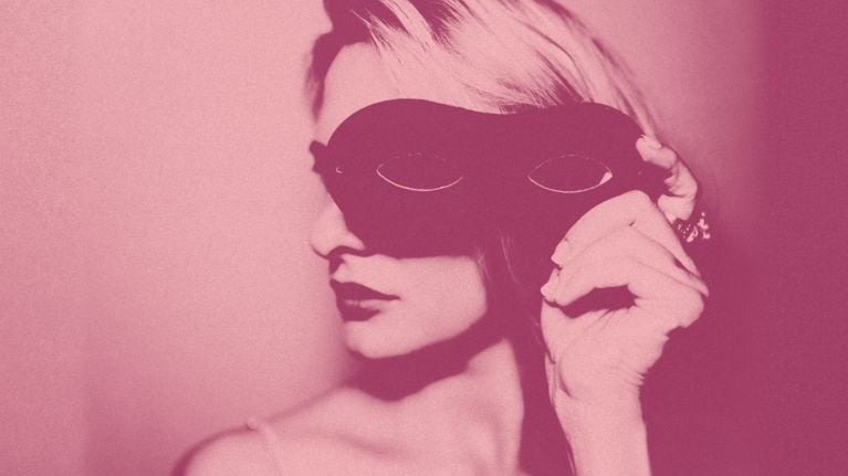 A photo of a woman holding a black masquerade eye mask at the side of her head, used in a post about one woman's first time at a sex club.