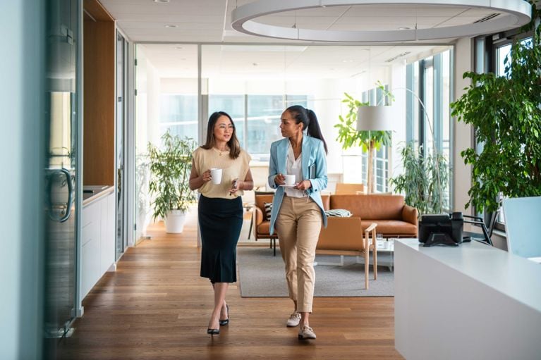 Two female coworkers holding cups of coffee and walking down a hall for a piece on sharing your salary in Canada
