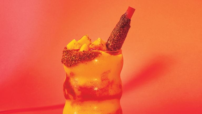 A wavy tall glass filled with mango slushy and stripes of chamoy sauce. It's dressed with chopped mango and a red straw.