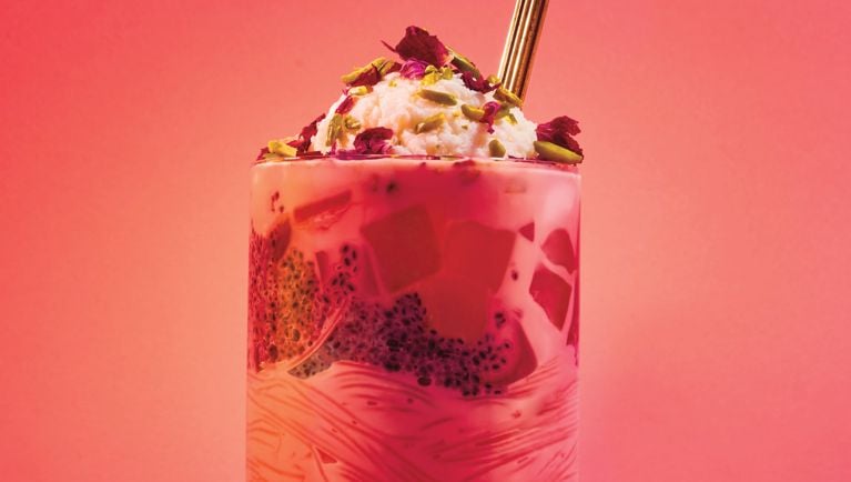 A tall highball glass filled with vermicelli noodles, sweet basil seeds, strawberry jello, ice cream, and pistachio rose petal garnishes.