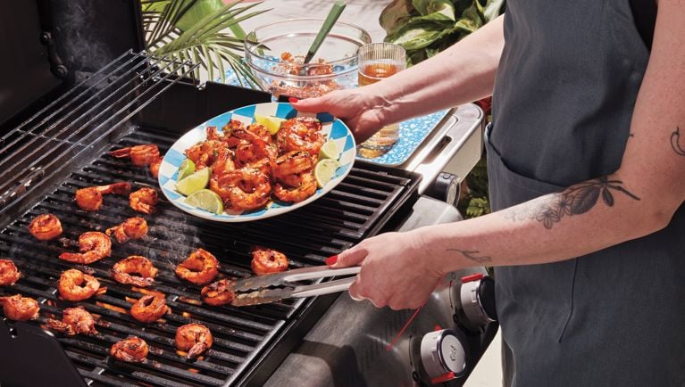 A woman is grilling shrimp on a Weber Spirit BBQ, and putting the shrimp on a bamboo platter.