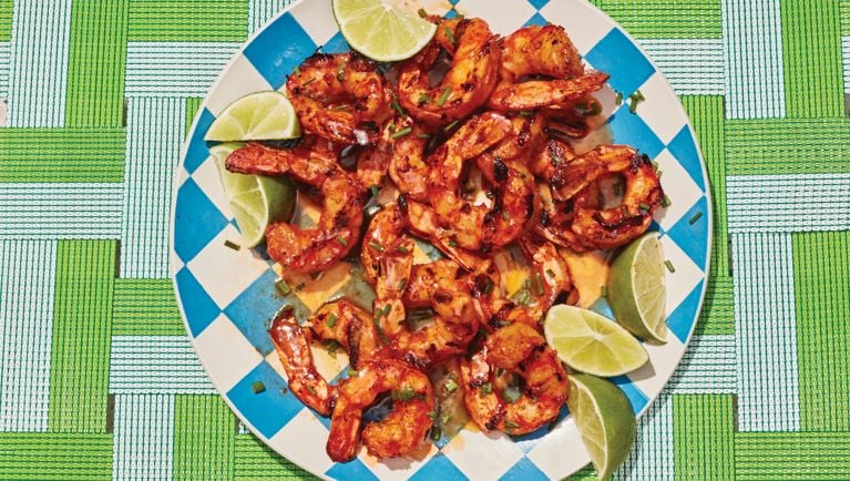 A large bamboo platter on a tablecloth loaded with freshly grilled hot honey shrimp. Slices of lime on the side of the plate.