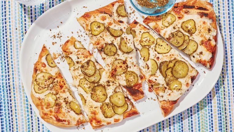 A freshly grilled flatbread spread with white sauce and loaded with sliced pickles and sumac is cut into triangles and arranged on an oval tray.