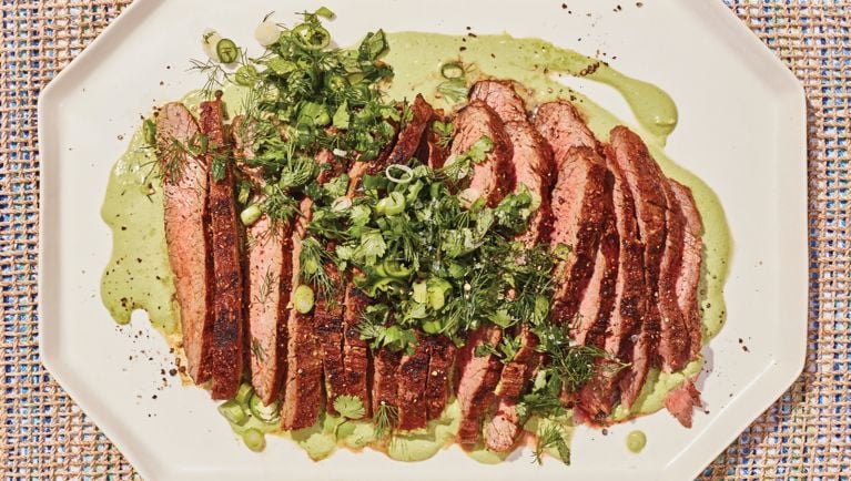 A freshly grilled flank steak is sliced thin and placed over a pool of bright green yogurt sauce, and topped with a pile of freshly chopped herbs.