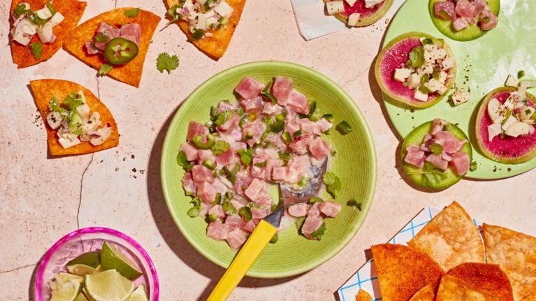A tuna ceviche with red onion and jalapeño sits un a shallow green bowl with a melamine fork. Some of the ceviche has been spooned onto tortilla chips on a separate plate.
