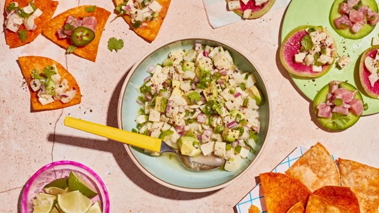 A vegan ceviche with hearts of palm, red onion and jalapeño sits un a shallow green bowl with a melamine fork. Some of the ceviche has been spooned onto tortilla chips on a separate plate.