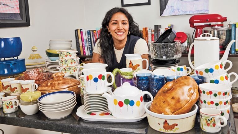 A woman sits behind a black granite counter that is completely loaded with piles of dishes, bowls, tea pots, cups, and soup tureens in varying styles and patterns.