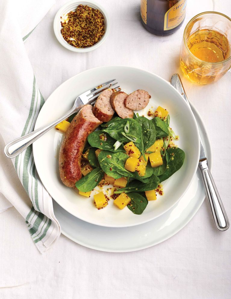 Bangers with a rutabaga sald on a white plate next to a glass of beer and a bowl of grainy mustard; the bangers are made with mild Italian sausage; the salad is made with cubed rutabaga; baby spinach; green onions and a whole-grain mustard vinaigrette for a post on an easy weeknight bangers with rutabaga salad recipe