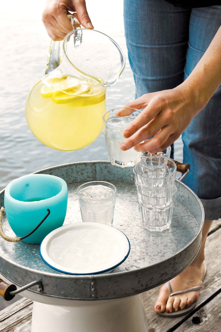 A pitcher of spiked lemonade, made with lemons, lemon juice, lemon slices, sugar and vodka being poured into a stack of glasses on a tray next to a candle and a bowl of ice on a dock with a woman's legs and feet in the background as she holds the pitcher for a post on an easy spiked lemonade recipe