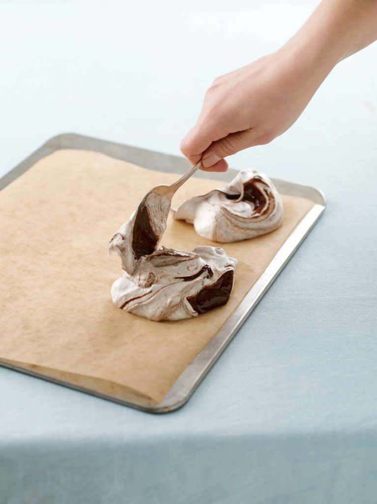 a hand dolloping two swirls of uncooked vanilla-chocolate meringues onto a flat baking sheet lined with parchment paper
