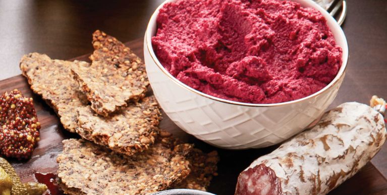 beet hummus in a white bowl next to seeded crackers and a sausage; the hummus is made with beets; olive oil; cauliflower; toasted walnuts; garlic; lemon juice; tahini; and coriander for a post on an easy, healthy beet hummus recipe for entertaining or dipping