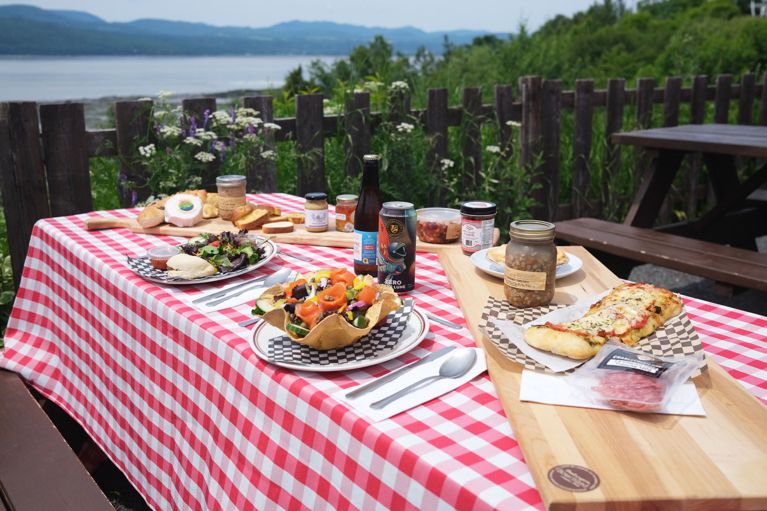 A picnic table spread with a red and white cocked tablecloth and laden with smoked salmon, fougases, drinks and more all over the St. Lawrence
