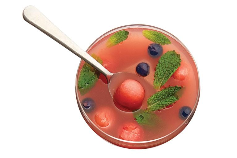 Chilled watermelon soup ina. bowl with a spoon resting in it; fresh mint leaves and blueberries and watermelon balls float in it. The soup is made with watermelon; sugar; fresh mint; white wine; and blueberries for a post on a refreshing watermelon soup recipe for dessert