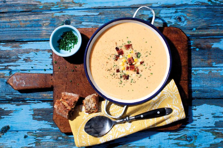 a bowl of creamy corn soup topped with bacon and chopped chives next to a piece of baguette. The corn soup is made with onion; olive oil; corn; garlic; chopped chipotle in adobo sauce; 35% cream; salt and chives and bacon for a garnish for a post on an easy, fast creamy corn soup recipe