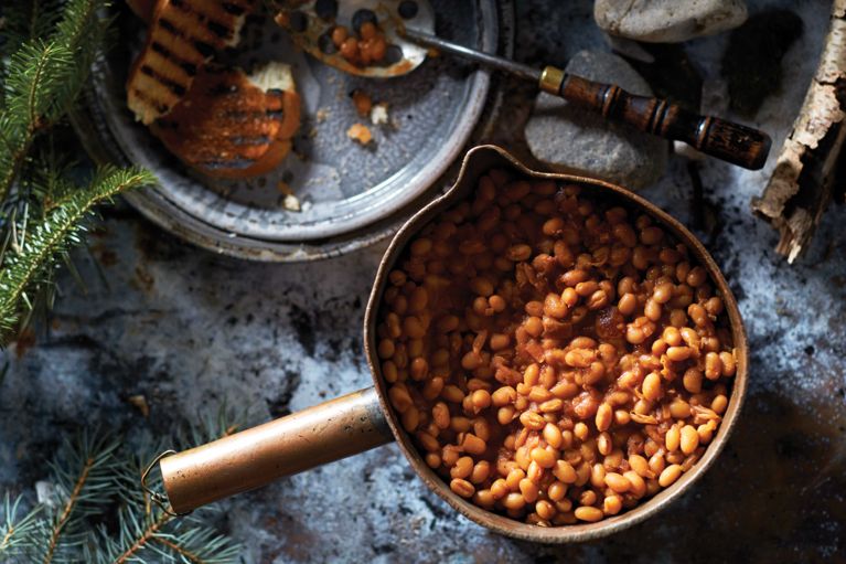 quick vegetarian baked beans with molasses and chipotle in a pot on a stone in an outdoorsy setting next to a ladle; the beans are made with onion; molasses; maple syrup; tomato paste; Dijon mustard; chipotle chili pepper powder; canned navy beans; and water for a post on a quick, easy vegetarian baked beans recipe