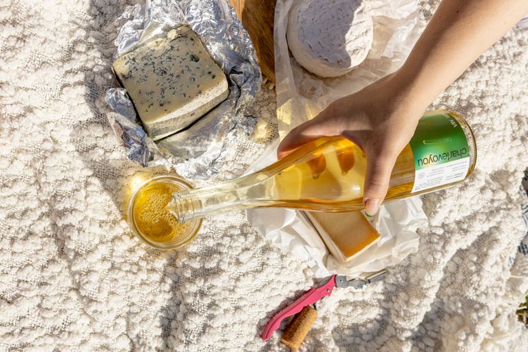 a hand holding a bottle of white wine next to cheese on a picnic blanket