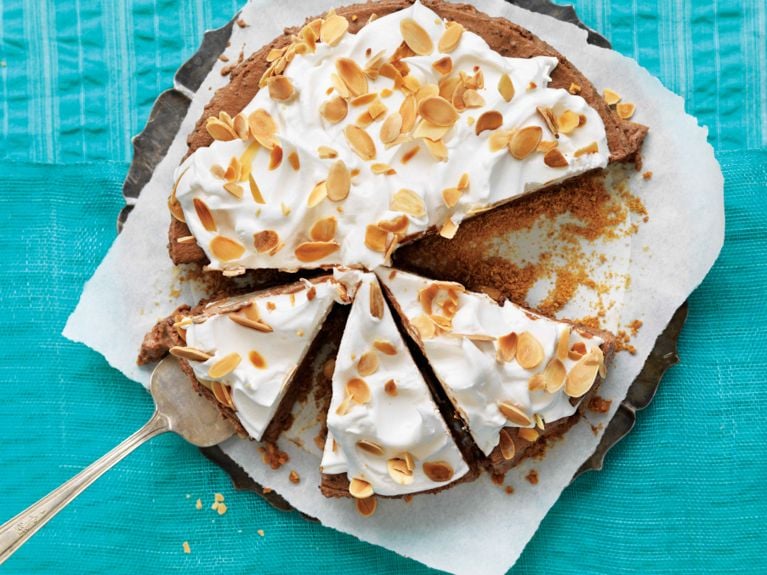 A frozen s'mores cake on a platter lined with parchment paper with a slice removed; the cake is made with graham-cracker crumbs; 35% cream; chopped bittersweet chocolate; and egg whites with a meringue topping topped with toasted sliced almonds for a post on a refreshing frozen s'mores cake recipe
