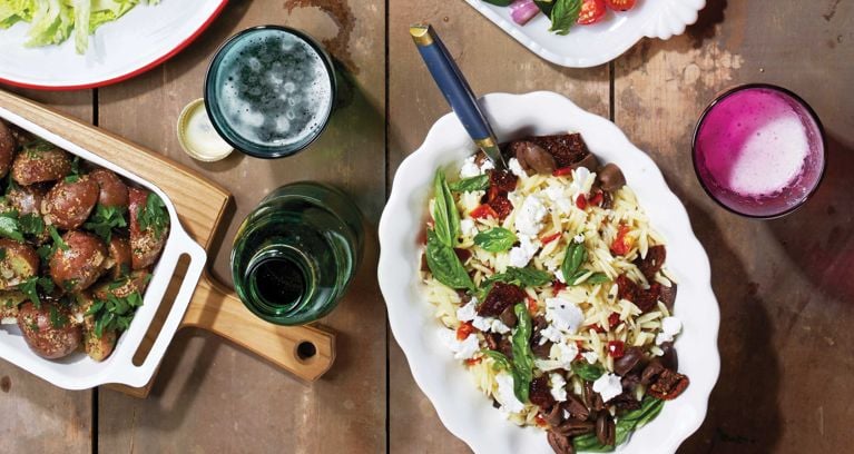 A greek goddess pasta salad on a white platter on a picnic table surrounded by summer dishes and glasses of beer; the salad is made with orzo; red wine vinegar; sugar; salt; extra-virgin olive oil; finely chopped sun-dried tomatoes; pitted kalamata olives; goat cheese and fresh basil