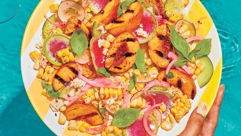 A Grilled Peach, Fresh Corn And Feta Salad on a yellow and white platter over a pool; the salad is made with red onion; white vinegar; honey; salt; limes; peaches; olive oil; corn; radishes; feta; chilli powder and basil for a post on an easy grilled corn and peach with feta salad recipe for summer entertaining
