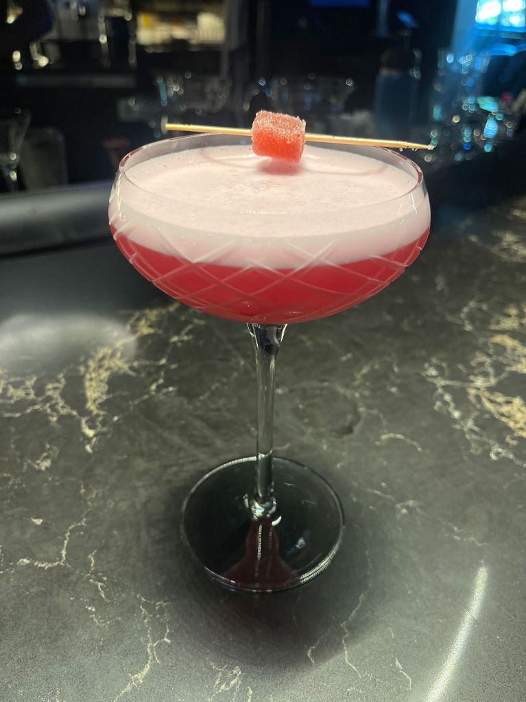 A pink cocktail with a frothy head in a coupe glass topped with a pink gummy on a toothpick