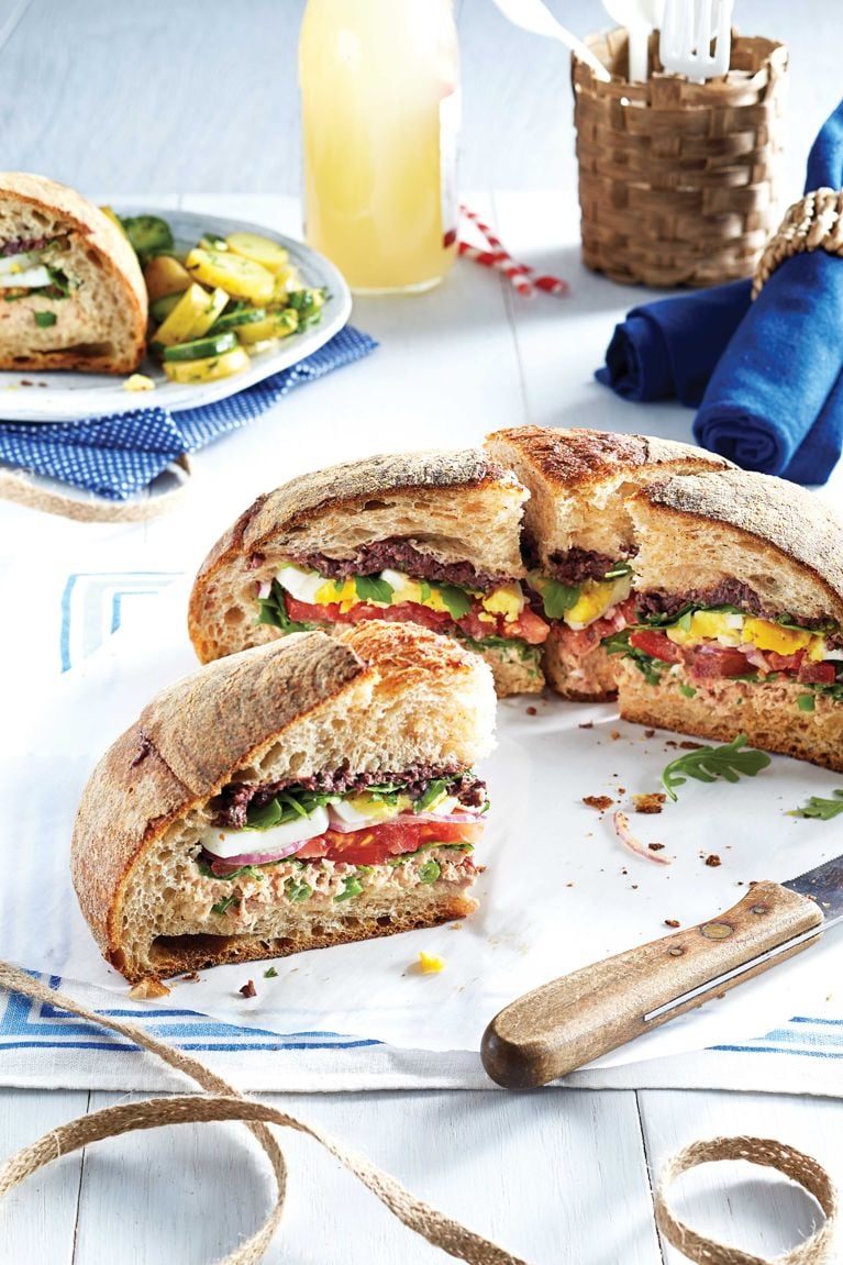 Niçoise muffuletta sandwich on a sheet of parchment paper next to a picnic spread; the sandwich is made with hard-boiled eggs; green beans; tuna; dijon mustard; mayonnaise; kalamata olives; anchovies; garlic; Italian bread; olive oil; arugula; tomato and red onion for a post on an easy nicoise muffuletta sandwich recipe for a picnic