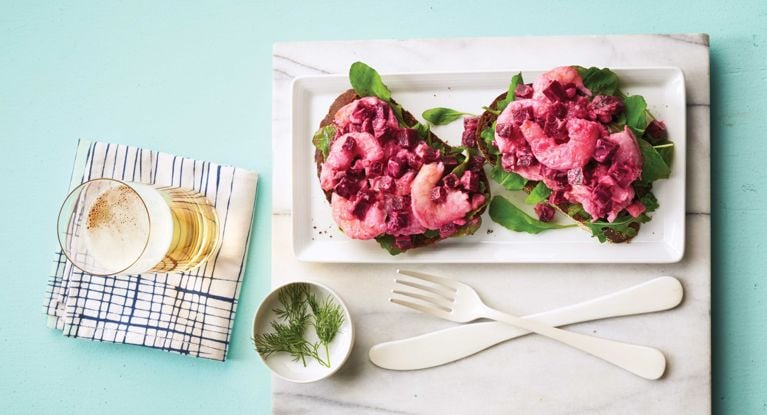 A nordic beet shrimp sandwich made with mayo; minced shallot; dijon mustard; sugar; pepper; chopped pickled baby beets; cooked shrimp; on rye bread with arugula next to a bowl of fresh dill and a glass of beer for a post on an easy nordic beet shrimp sandwich recipe