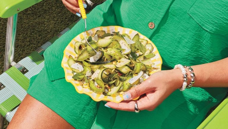 A woman in a green jumper set sit on a chair and eats zucchini and mizithra salad from a yellow scalloped plate for a post on a delicious, easy zucchini salad recipe for summer