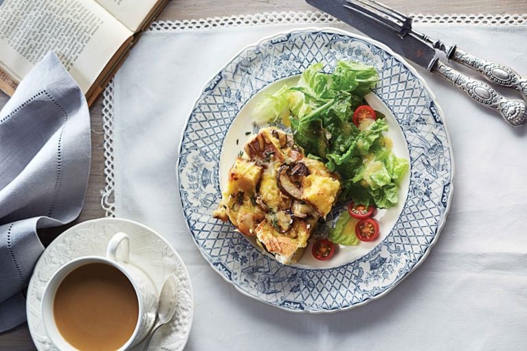 Slow Cooker Cheese And Mushroom Strata on a blue and white plate next to a green salad; the Strata is made with leftover stale bread; sliced cremini mushrooms; olive oil; gruyere or Jarlsberg cheese; eggs; milk; thyme; dijon mustard and salt for a post on an easy slow cooker mushroom and cheese strata recipe for breakfast or for summer