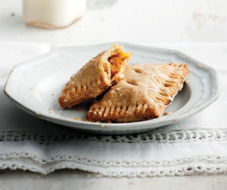 S'mores hand pies on a white platter; individual pies filled with mini marshmallows and chocolate with a maple glaze for a post on a s'mores hand pies recipe