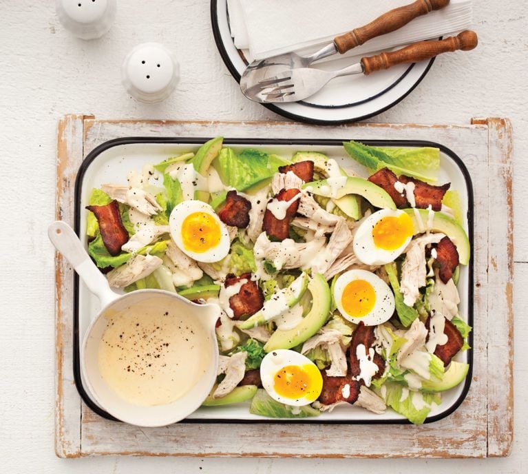 A summer club salad on a rectangular tray ready to be eaten. The salad is made with soft-boiled eggs; skinless, boneless chicken breasts; bacon; mayonnaise; Dijon; white vinegar; avocado next to a pot full of dressing for a post on an easy weeknight summer club salad recipe