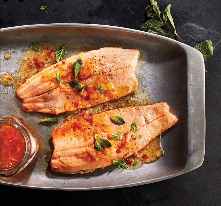 Sweet and spicy grilled rainbow trout on a platter topped with leaves of mint and next to a jar of marmalade-sriracha sauce for a post on an easy Sweet and spicy grilled rainbow trout recipe for summer