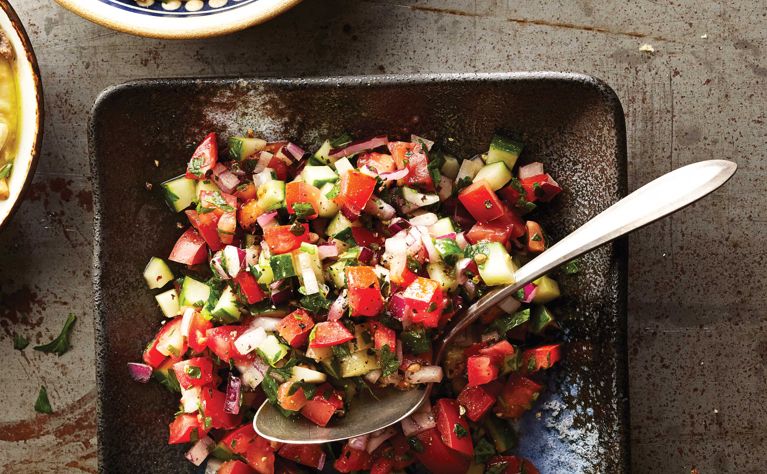 Tomato and cucumber chopped salad on a plate with a spoon resting in it; the salad also contains red onion; salt; olive oil; lemon juice and parsley for a post on an easy tomato cucumber chopped salad recipe