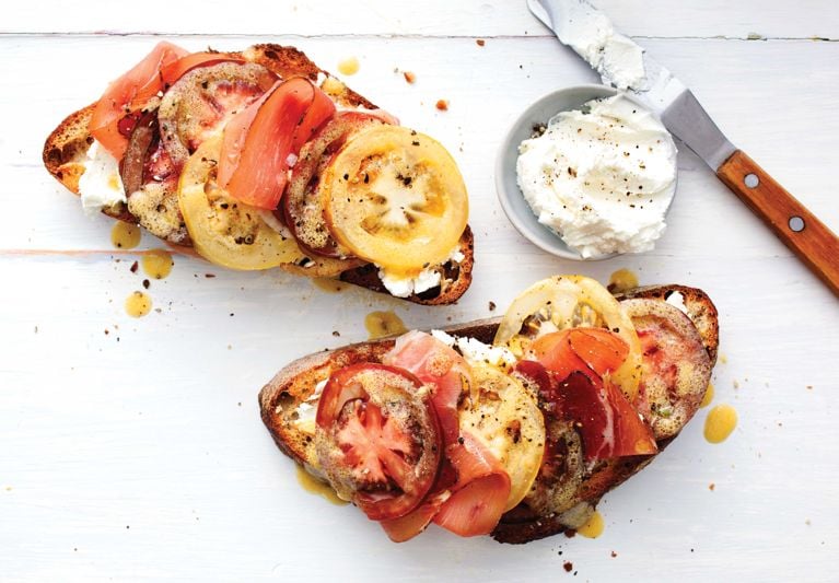 a tomato and prosciutto open-faced sandwich on rustic bread next to a small bowl of goat cheese with a spreading knife; the sandwiches are made with red beefsteak tomato, sliced into 1/4-in. rounds; yellow beefsteak tomatoes; white balsamic vinegar; olive oil; honey; dijon mustard; garlic; sourdough bread; goat cheese; prosciutto and pepper for a simple open-faced summer tomato sandwich recipe