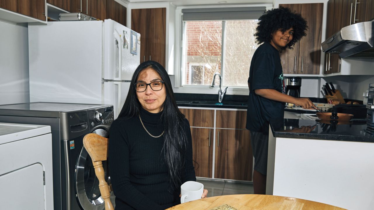 Sheri Pranteau sits at her kitchen table while her son, Neville, prepares food on the stove.