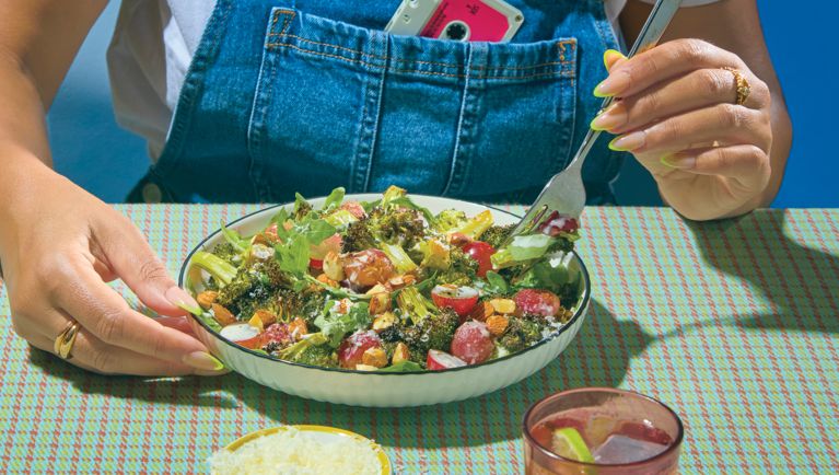 A large shallow bowl of broccoli and grape salad with someone sitting at a table and putting a fork into the salad.