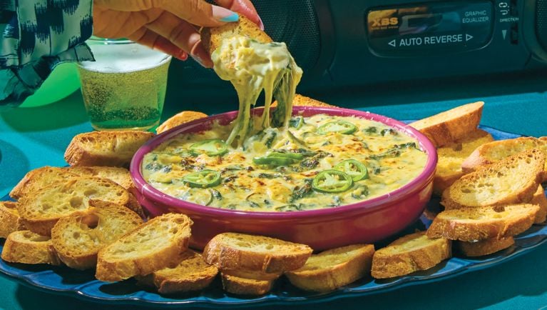 A wide pink bowl of cheese and artichoke dip surrounded by sliced baguette. Someone is dipping a piece of bread into the dip.