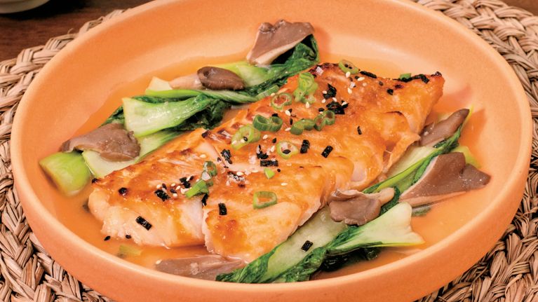 A slice of glazed cod on a pink plate with large pieces of bok choy and mushrooms surrounding it.