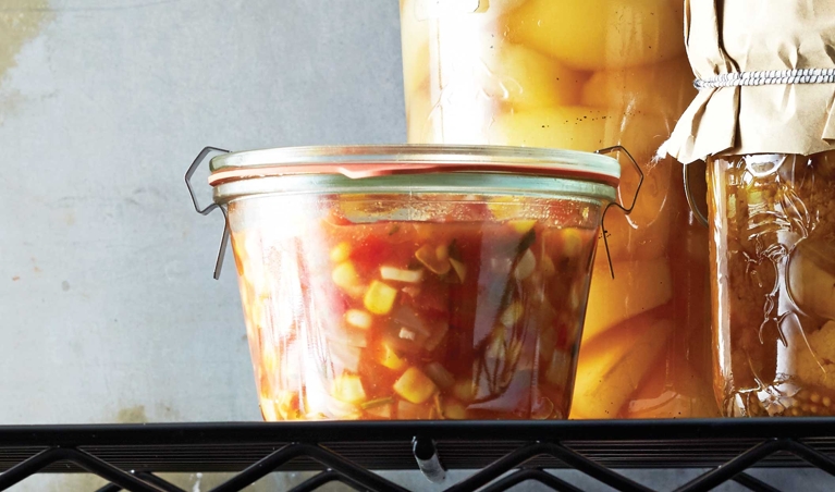 A series of jars of preserves and salsas on black wire racks, including a container of fresh summer salsa made with tomatoes; diced onion; corn kernels; diced red bell peppers; white vinegar; garlic; minced, seeded jalapeño; cilantro; sugar; cumin and salt for a post on an easy fresh summer salsa recipe
