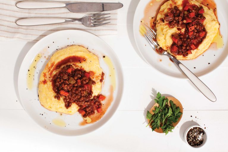 Two plates with lentil rags over polenta, next to bowls of fresh parsley and cracked black pepper for a post on an easy two-for-one lentil rags over polenta recipe