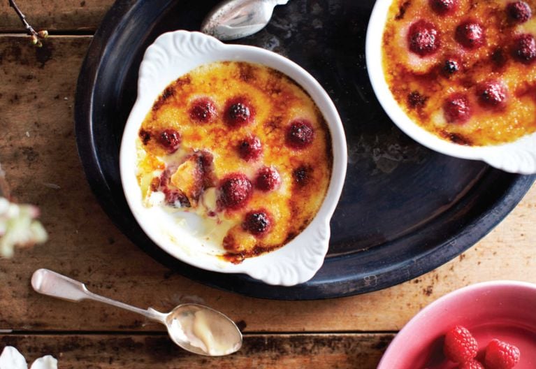 Two white ramekins full of Raspberry And Basil Créme Brûlée on a try, next to a spoon and a bowl of raspberries; the creme brûlée is made with fresh raspberries; egg yolks; sugar; 5% cream; and vanilla for a post on a raspberry basil creme brûlée recipe