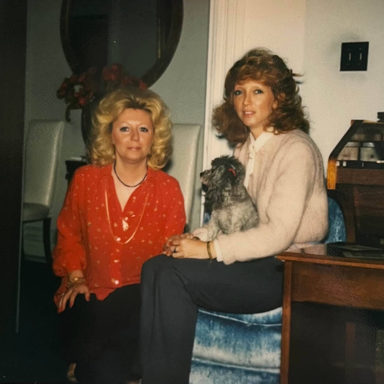 A phooto of a blonde woman in a red blouse and a brunette woman in a beige sweater with a grey poodle on her lap.