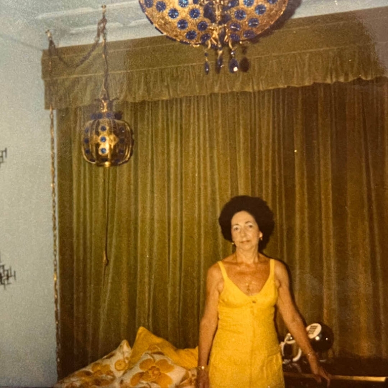 A photo of a woman in a yellow dress in a living room with ornate lamps and gold drapes.